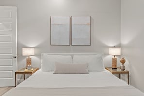 Un dormitorio con una cama, dos mesitas de noche, una puerta y dos marcos de cuadros en la pared.
