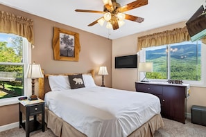 A bedroom with a bed, bedside lamps, a ceiling fan, a TV, a dresser, and a window with a view of greenery. - Mountain-View Condo • Shared Pool + Hot Tub • Near Gatlinburg | Lazy Bear (Gatlinburg)