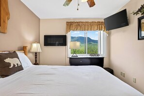 A hotel room with a bed, a desk with a lamp, a TV mounted on the wall, and a window with a view of mountains. - Mountain-View Condo • Shared Pool + Hot Tub • Near Gatlinburg | Lazy Bear (Gatlinburg)