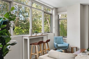 Ein Zimmer mit großen Fenstern, einem blauen Sessel, einem weißen Sofa und einem hölzernen Hocker.