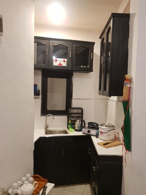 A compact kitchen with a sink, a stove, and a microwave. There is a cabinet above the sink and a shelf with kitchen items.