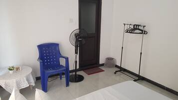 A room with a bed, a blue chair, a standing fan, a clothes drying rack, and a small table with a plant.