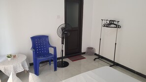 Una habitación con una cama, una silla azul, un ventilador de pie, un tendedero y una pequeña mesa con una planta.