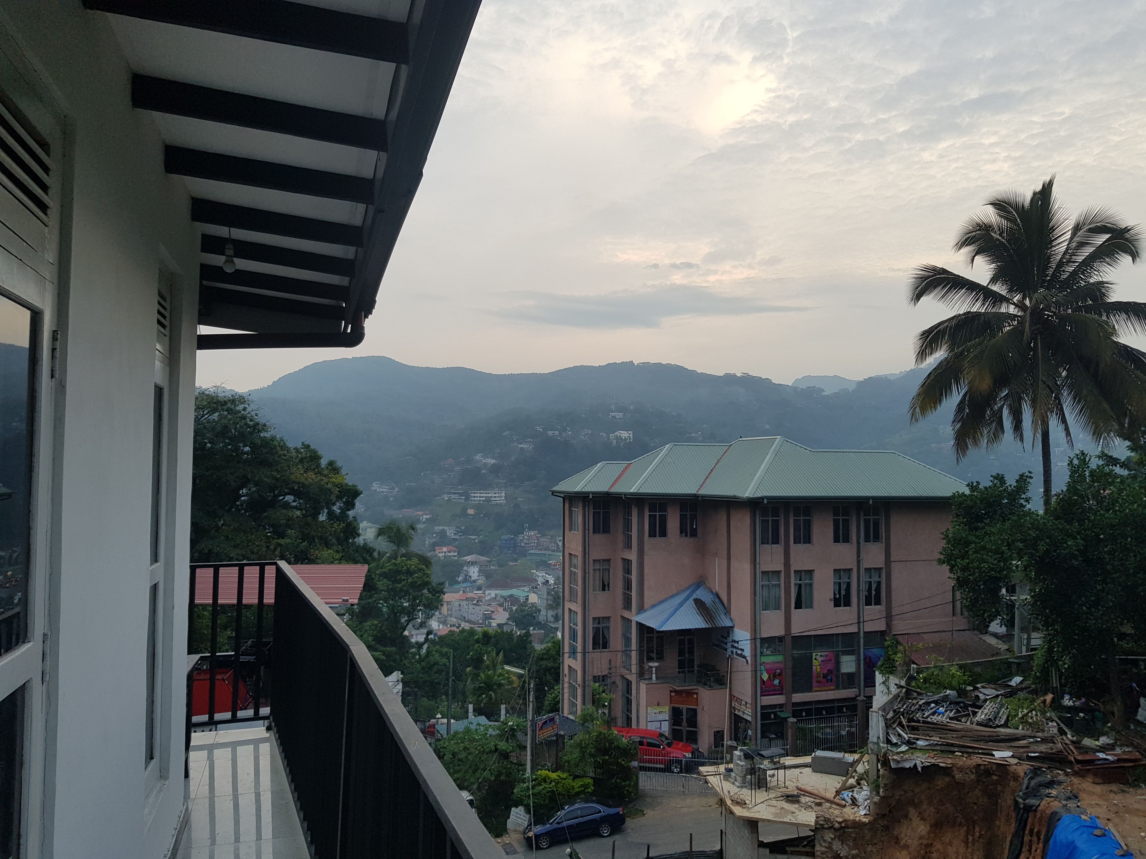 A multi-story building with a flat roof, a balcony, and a view of a hilly landscape in the background.