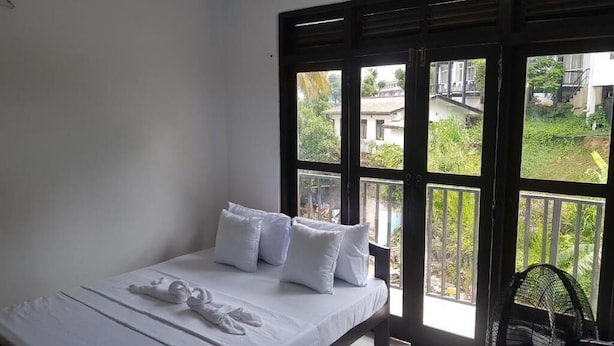 A bedroom with a bed, pillows, and a window with a view of greenery and buildings. - Shalom View Homestay  (Kandy)
