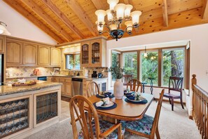 A spacious kitchen with wooden cabinets, a dining area with a table set for four, and a large window offering a view of the outdoors. - Alpine Chalet + Decks + Game Room | Near Ski + Lake | Stillwood (Stateline)