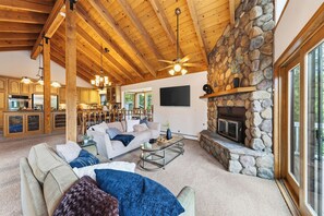 A spacious living area with a stone fireplace, wooden ceiling beams, and a modern kitchen in the background. - Alpine Chalet + Decks + Game Room | Near Ski + Lake | Stillwood (Stateline)