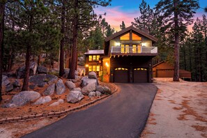 A two-story cabin with a garage, surrounded by pine trees and a driveway leading to the entrance. - Alpine Chalet + Decks + Game Room | Near Ski + Lake | Stillwood (Stateline)
