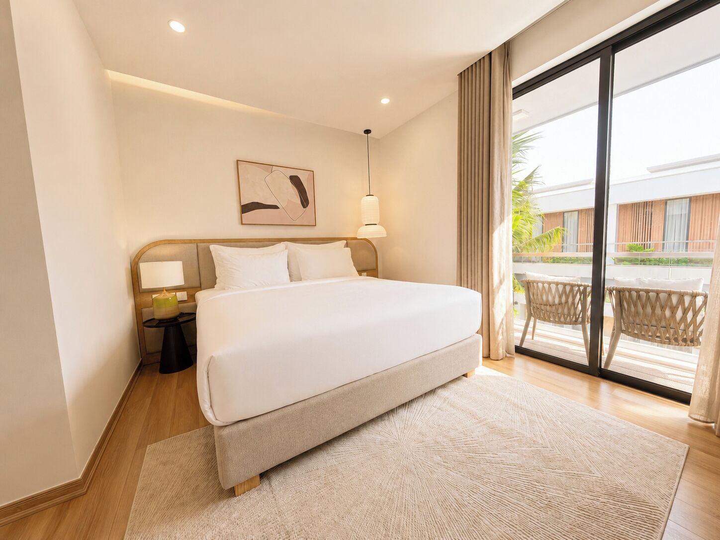 Habitación de hotel con una cama grande, una mesita de noche, una lámpara de pared y vistas al balcón exterior.