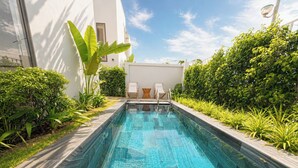 Una piscina con superficie de agua azul cristalina, rodeada de exuberante vegetación y un edificio blanco al fondo.