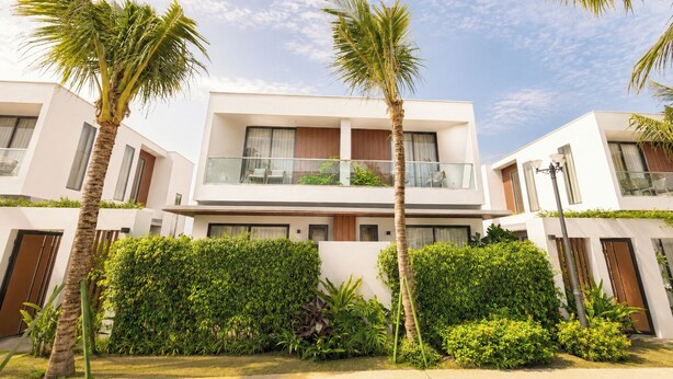 Un edificio moderno de dos pisos con balcones, rodeado de palmeras y abundante vegetación cuidada.
