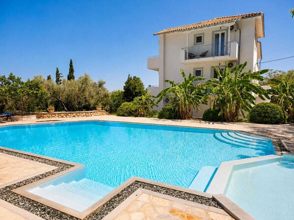 A swimming pool with a clear blue water, surrounded by a stone deck, and a two-story building with a balcony in the background. - Villa Irini Ocean View 2-bedroom apartment with AC in charming Fryni (Fryni)