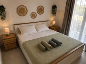 A neatly made bed with a green blanket and rolled towels, two bedside tables with lamps, and decorative wall hangings. - Villa Irini Ocean View 2-bedroom apartment with AC in charming Fryni (Fryni)