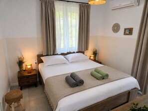 A neatly made bed with a beige bedspread and pillows, a bedside lamp, a small wooden nightstand with a lamp, and a wicker stool. - Villa Irini Ocean View 2-bedroom apartment with AC in charming Fryni (Fryni)