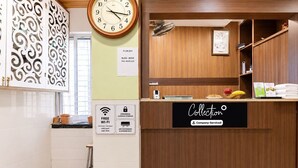 Ein Hotel-Rezeptionsbereich mit einem Holztresen, einer Uhr und einem Schild, das Annehmlichkeiten wie kostenloses WLAN, Gepäckaufbewahrung durch den Concierge und Klimaanlage anzeigt.