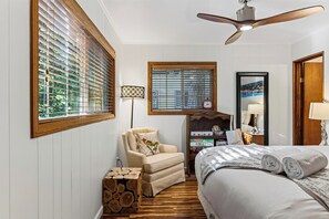 A bedroom with a bed, a chair, a wooden side table, a bookshelf, and a ceiling fan. - Forest Views + Firepit Gazebo + Deck Dining | Near Village | Wilderness Lodge (Lake Arrowhead)