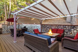 An outdoor patio with a stone wall, wicker furniture, a fire feature, and a pergola. - Forest Views + Firepit Gazebo + Deck Dining | Near Village | Wilderness Lodge (Lake Arrowhead)