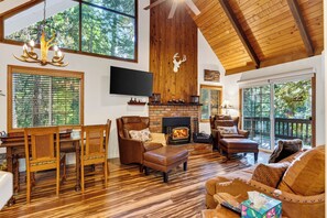 A spacious living room with a fireplace, wooden furniture, and large windows offering a view of the outdoors. - Forest Views + Firepit Gazebo + Deck Dining | Near Village | Wilderness Lodge (Lake Arrowhead)