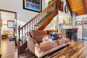 A spacious living room with a wooden staircase, a sofa with decorative pillows, and a dining area with a view. - Forest Views + Firepit Gazebo + Deck Dining | Near Village | Wilderness Lodge (Lake Arrowhead)