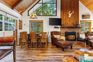A spacious living room with a fireplace, wooden furniture, and large windows. - Forest Views + Firepit Gazebo + Deck Dining | Near Village | Wilderness Lodge (Lake Arrowhead)
