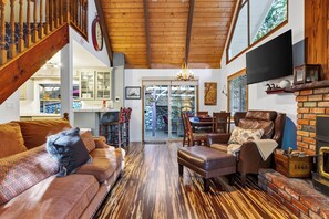A cozy living room with a fireplace, leather sofas, and a dining area with a stone wall backdrop. - Forest Views + Firepit Gazebo + Deck Dining | Near Village | Wilderness Lodge (Lake Arrowhead)