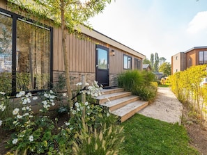 Una casa moderna con fachada de madera, puerta negra y jardín con flores blancas y follaje verde.