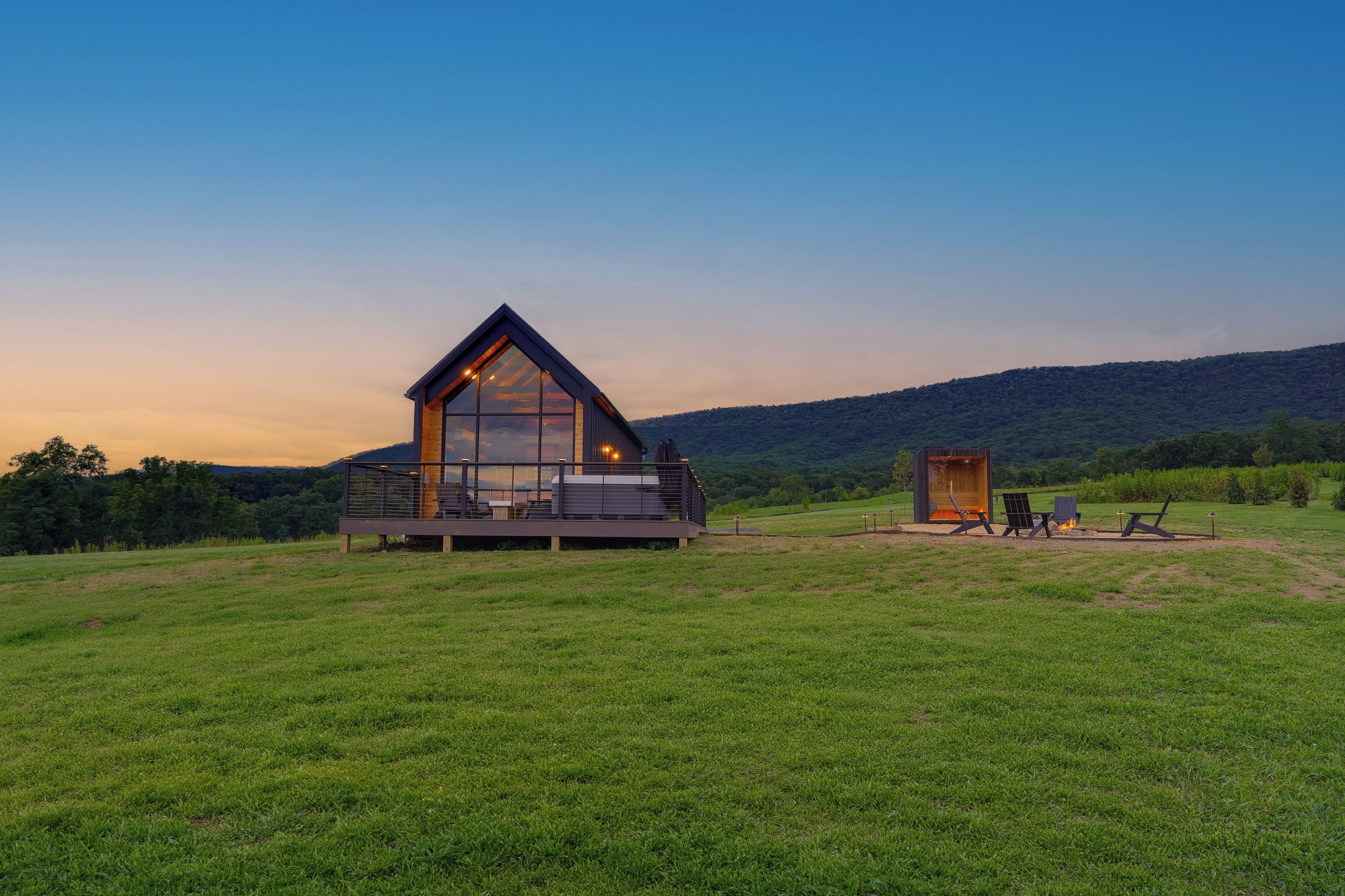 A modern cabin with a large glass facade, a deck, and a small adjacent structure, set in a grassy field with a forested hill in the background.