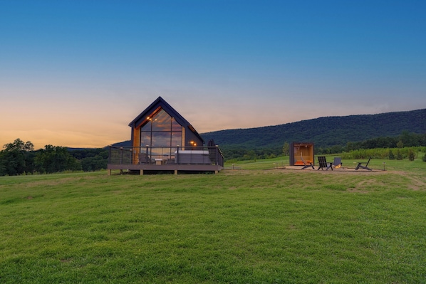 A modern cabin with a large glass facade, a deck, and a small adjacent structure, set in a grassy field with a forested hill in the background.