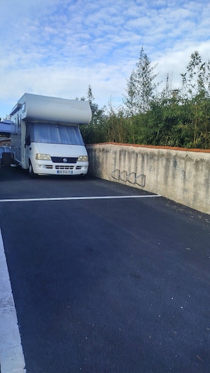 Un camping-car blanc est garé sur une surface goudronnée, avec un mur en béton et de la végétation en arrière-plan.