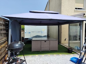 Un espace de cuisson extérieur abrité, équipé d’un barbecue et d’une cuisinière portative, d’une porte coulissante en verre et d’un petit jardin avec du gazon synthétique et du gravier.