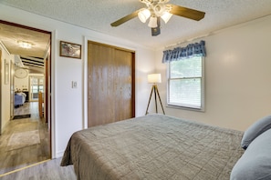 Quarto com cama, ventoinha no teto, janela com persianas e porta que dá para outro cômodo.