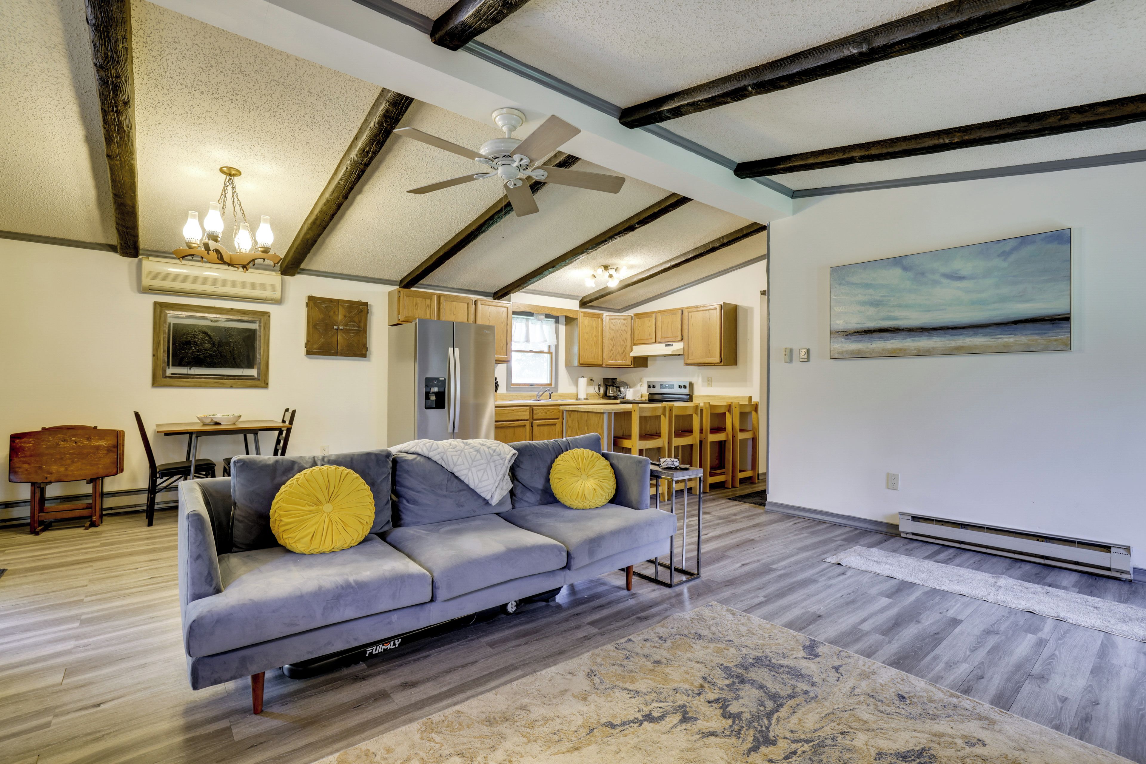 A modern living room with a gray sofa, wooden beams, a ceiling fan, and a kitchen in the background.