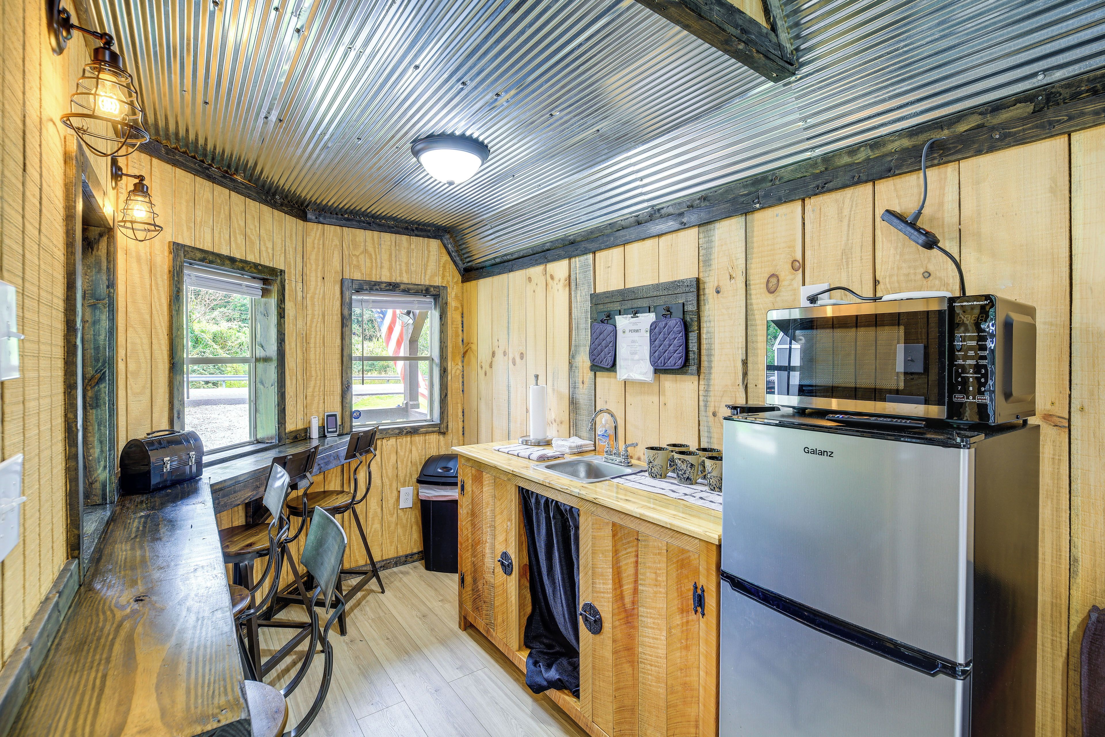 A compact kitchen with a stainless steel refrigerator, a microwave, and a sink. There are wooden walls and a window with an American flag.