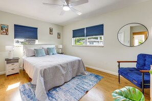 Ein Schlafzimmer mit Bett, Nachttischen, Deckenventilator, einem Fenster mit Jalousien, einem Spiegel und einem Sessel.