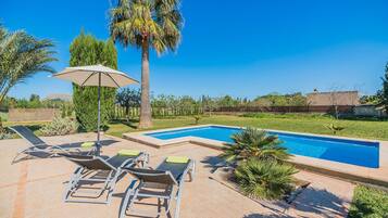 A swimming pool with lounge chairs and an umbrella, surrounded by greenery and a clear sky.