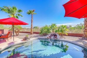 A swimming pool with a waterfall feature, surrounded by palm trees and red umbrellas. - Resort-Style Goodyear Home: Swim, Soak & Play! (Goodyear)