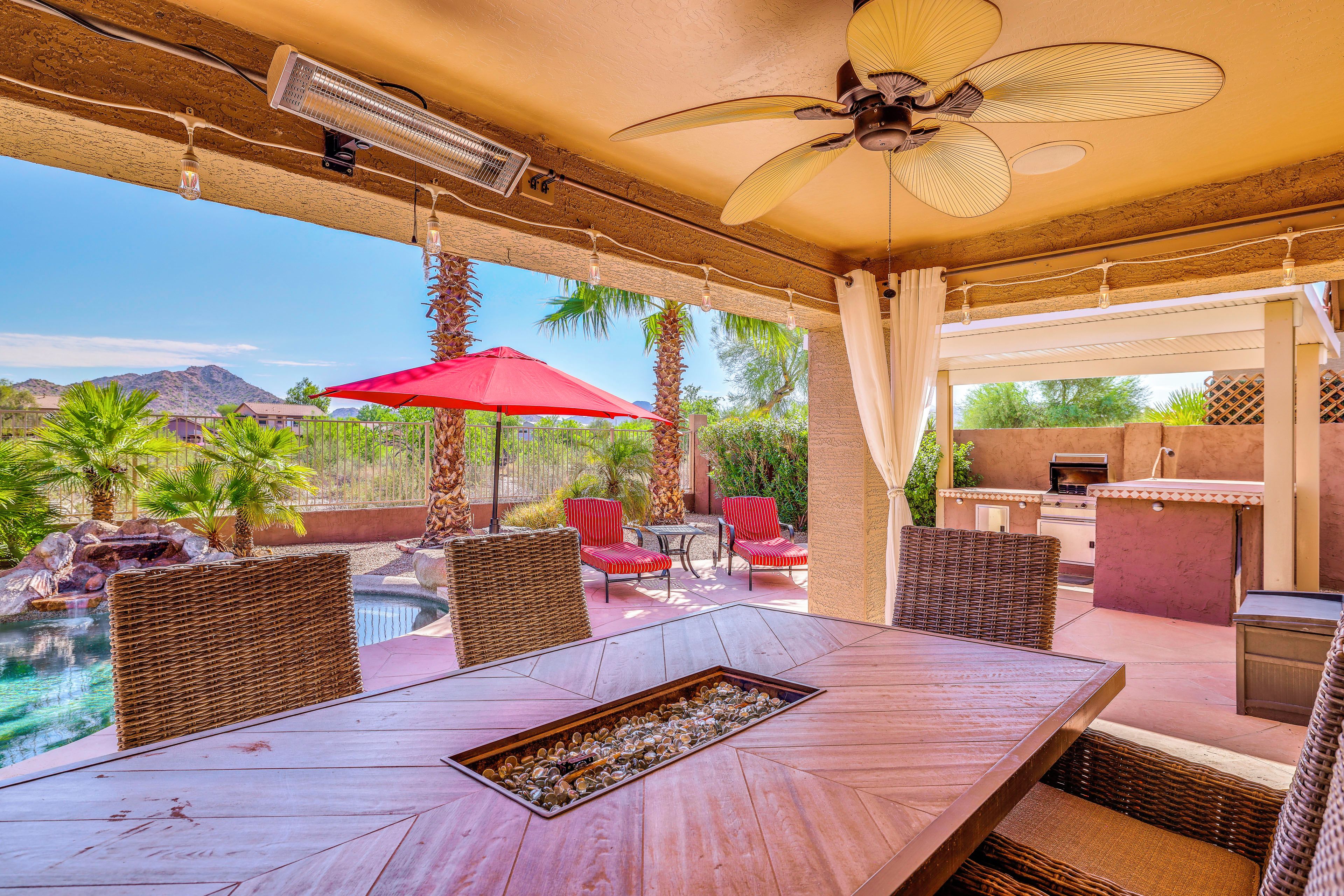 An outdoor patio with a dining table, wicker chairs, a red umbrella, and a view of a pool and mountains.