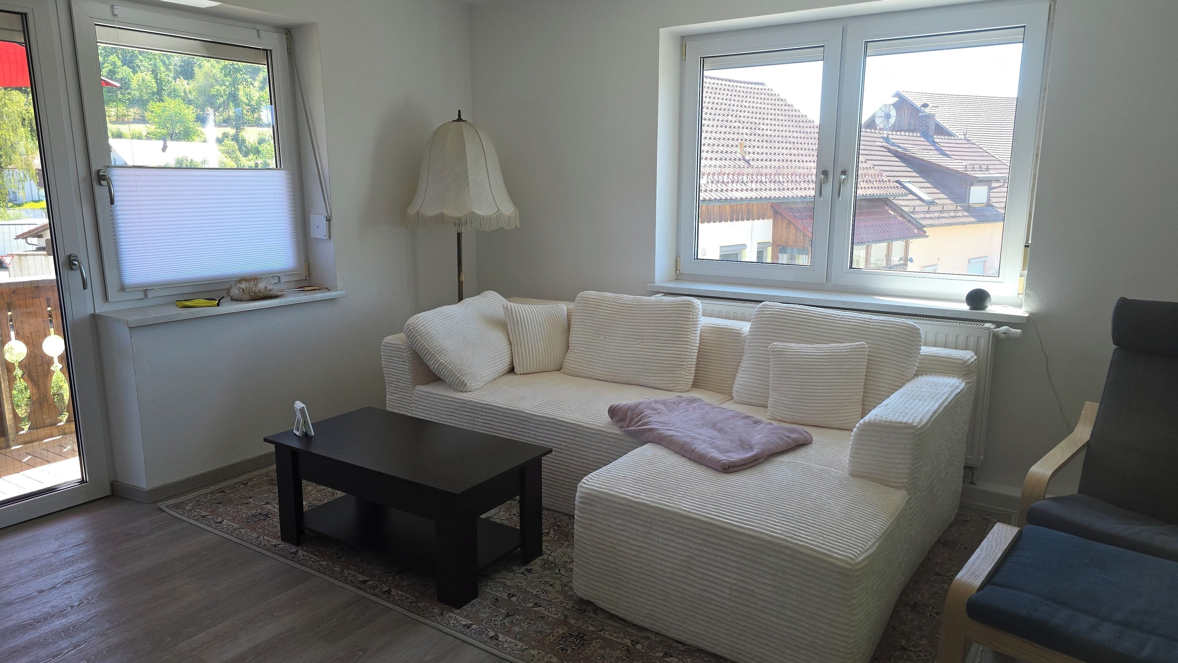 Ein Wohnzimmer mit einem weißen Sofa, einem kleinen schwarzen Tisch und einer Lampe. Fenster bieten Blick auf Gebäude und Bäume.