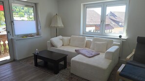 Sala de estar con un sofá blanco, una pequeña mesa negra y una lámpara. Se ven edificios y árboles a través de las ventanas.