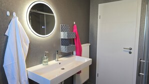 A bathroom with a white sink, a mirror with a light, a towel rack with a white towel, a pink towel, and a door. - Vrbo Property (Cham)