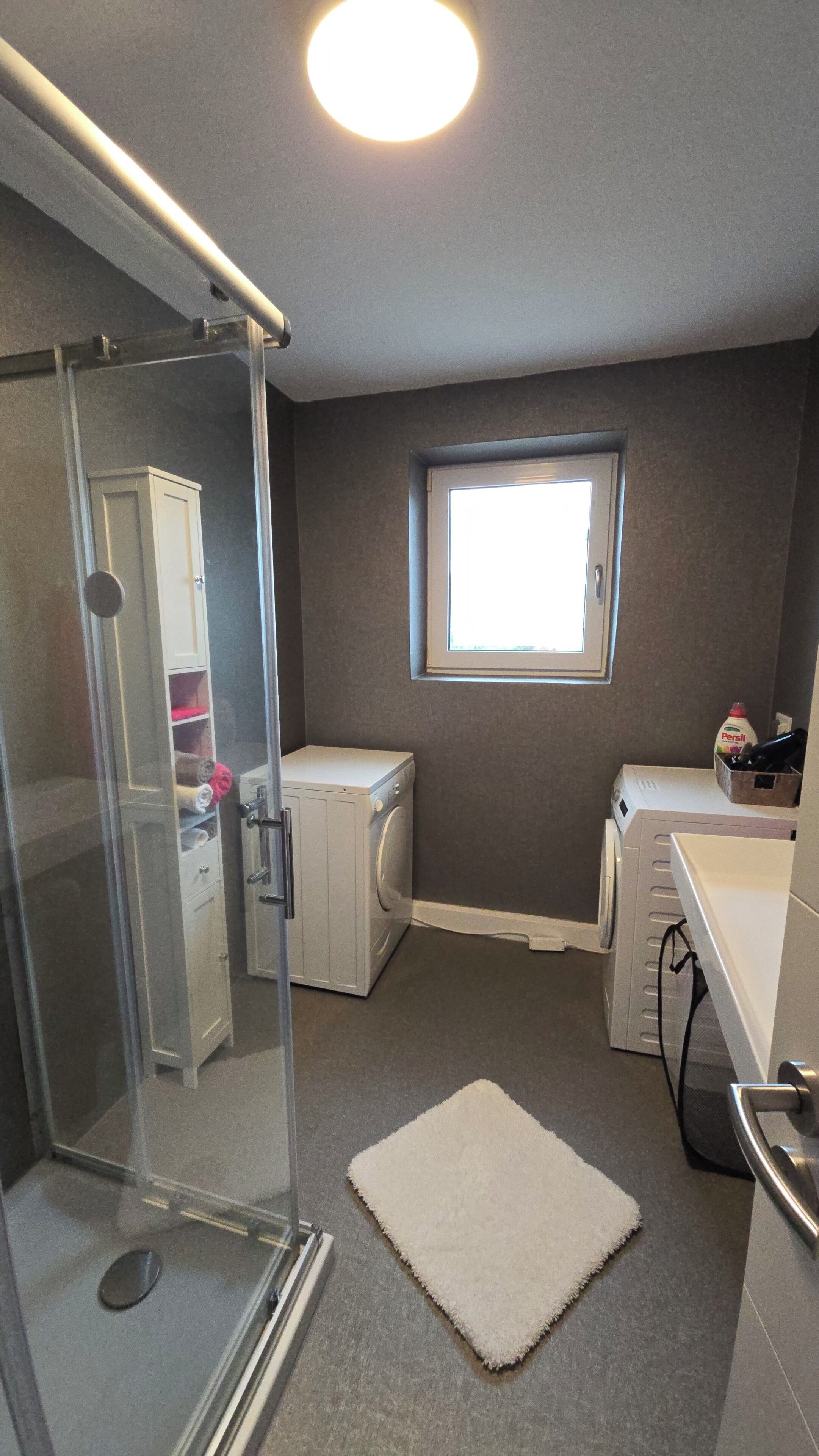 Ein kleines Badezimmer mit Duschkabine aus Glas, integrierter Waschmaschine und Trockner, einem kleinen Fenster und einem weißen Teppich auf dem Boden.