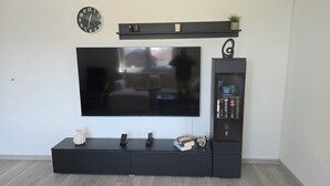 A modern living room with a flat-screen TV mounted on a black entertainment unit, a small shelf with decorative items, and a glass-fronted bookcase with books and decorative objects.