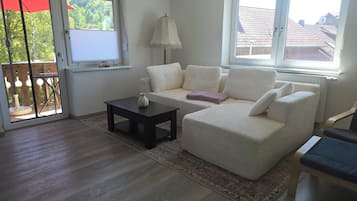 A living room with a sofa, a small table, and a lamp. There is a view of the outside through a window.
