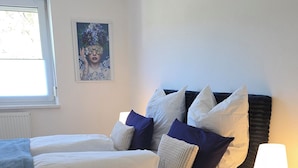 A bedroom with a bed, pillows, a bedside table with a lamp, a window, and a wall art piece.
