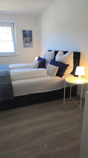 Un dormitorio con cama, almohadas, una mesita de noche con lámpara, una ventana y una obra de arte en la pared.