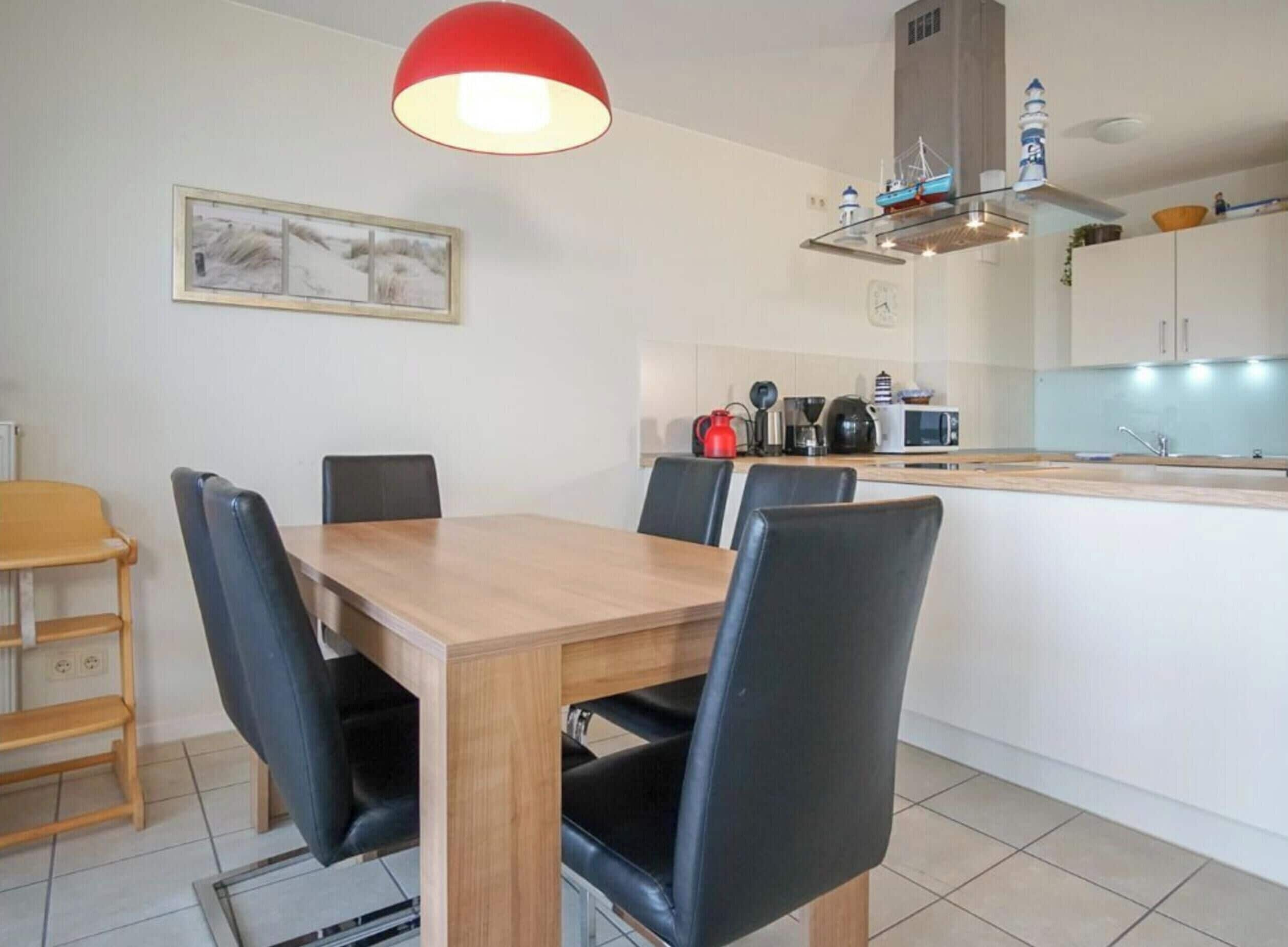 Un comedor con mesa de madera y sillas negras, una lámpara colgante roja, y una cocina con microondas y hervidor.