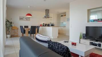 Una sala moderna con sofá, televisor y una mesita de centro. Se observa un comedor con sillas y una mesa, y una cocina al fondo.