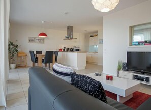Uma sala de estar moderna com sofá, televisão e mesa de centro. Há uma área de jantar com cadeiras e mesa, e uma cozinha ao fundo.