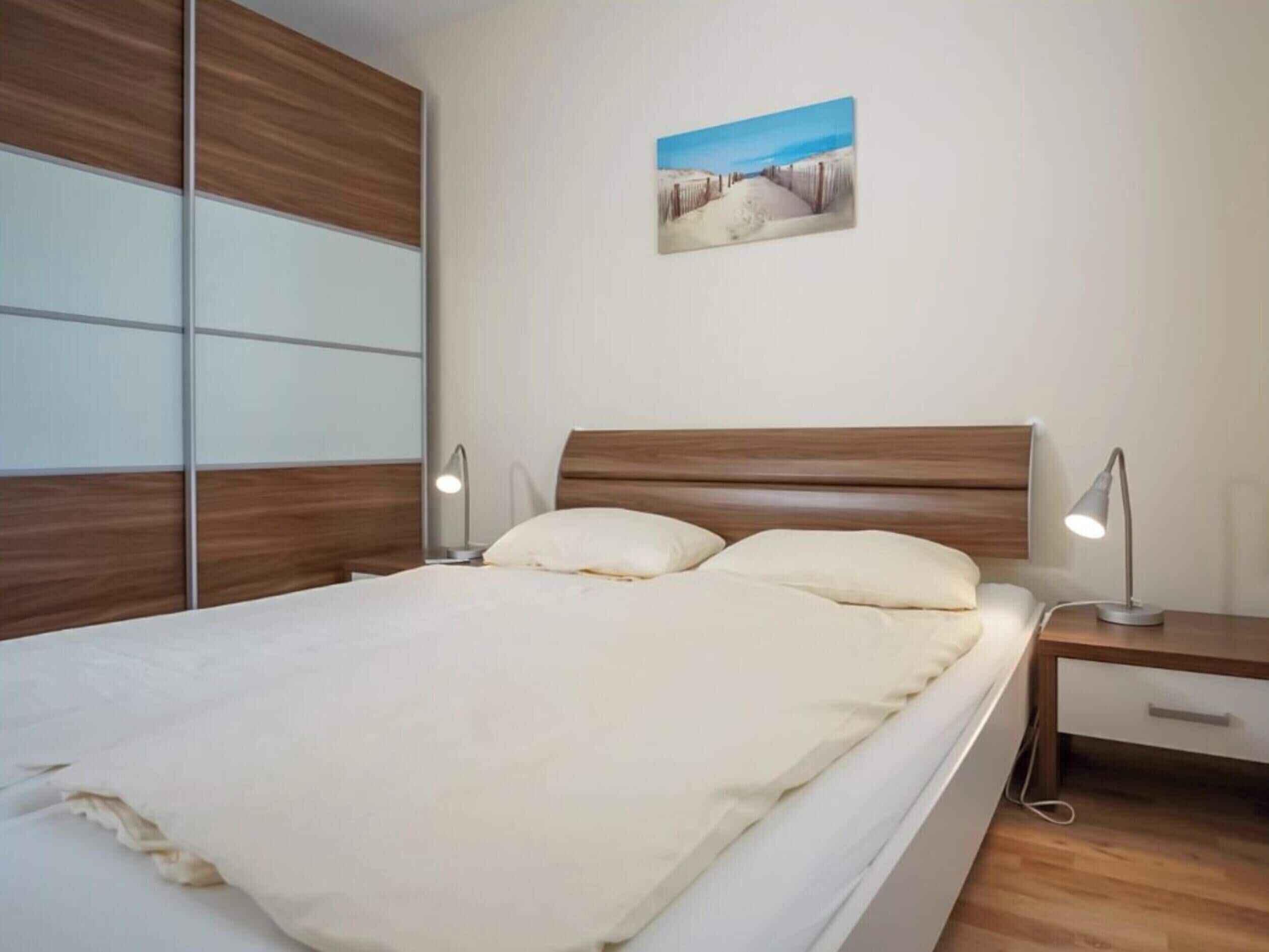Un dormitorio con cabecera de madera, cama con ropa de cama blanca, dos mesitas de noche con lámparas y un cuadro colgado en la pared.