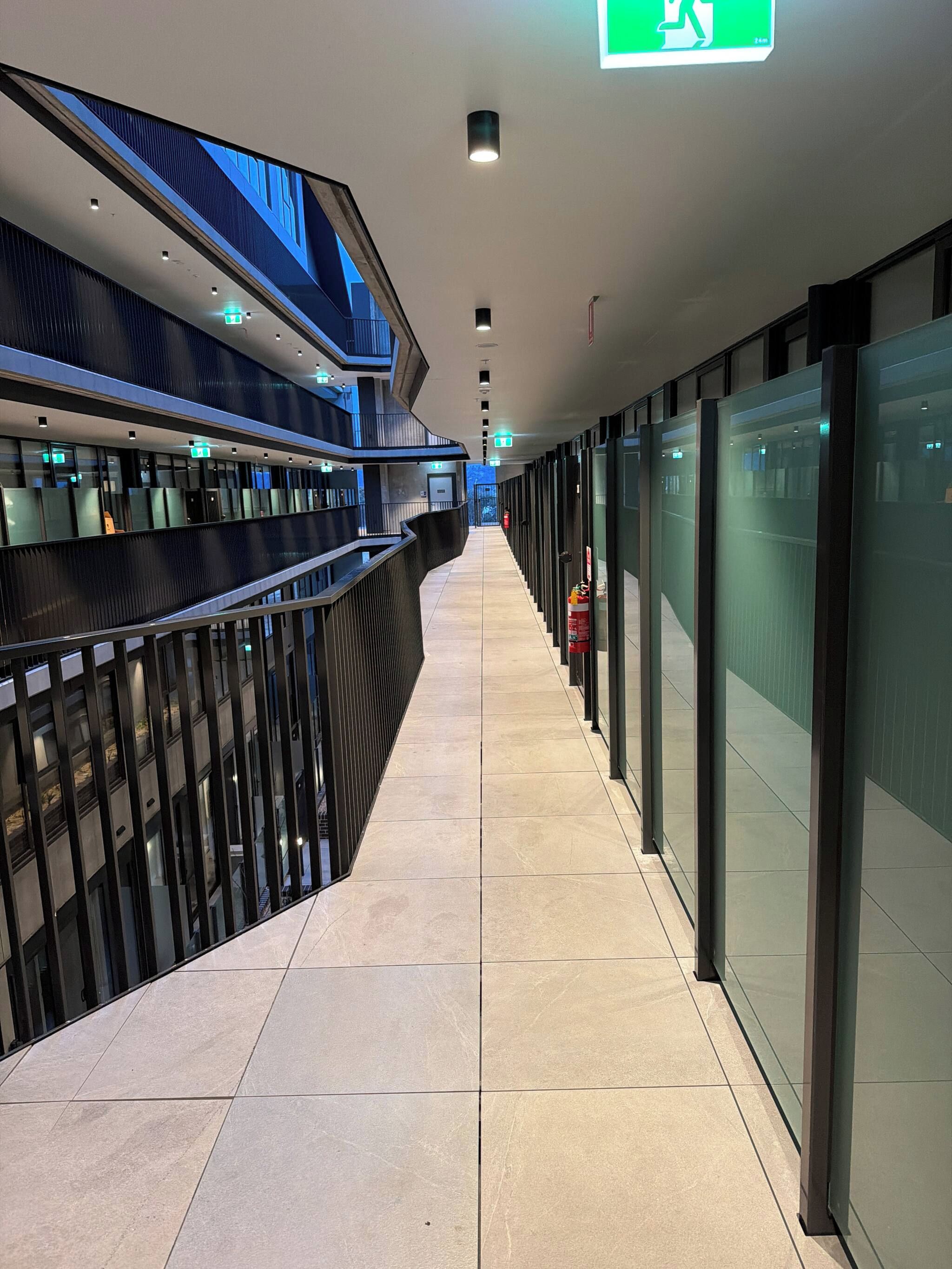 A corridor with glass walls, emergency exit signs, and a tiled floor.